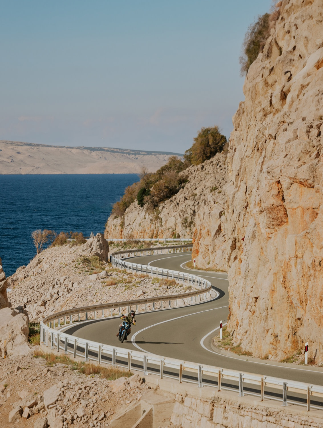 A Verge electric motorcycle rides on a serpentine-like mountain road by the seaside on a sunny day with cliffs alongside the road.
