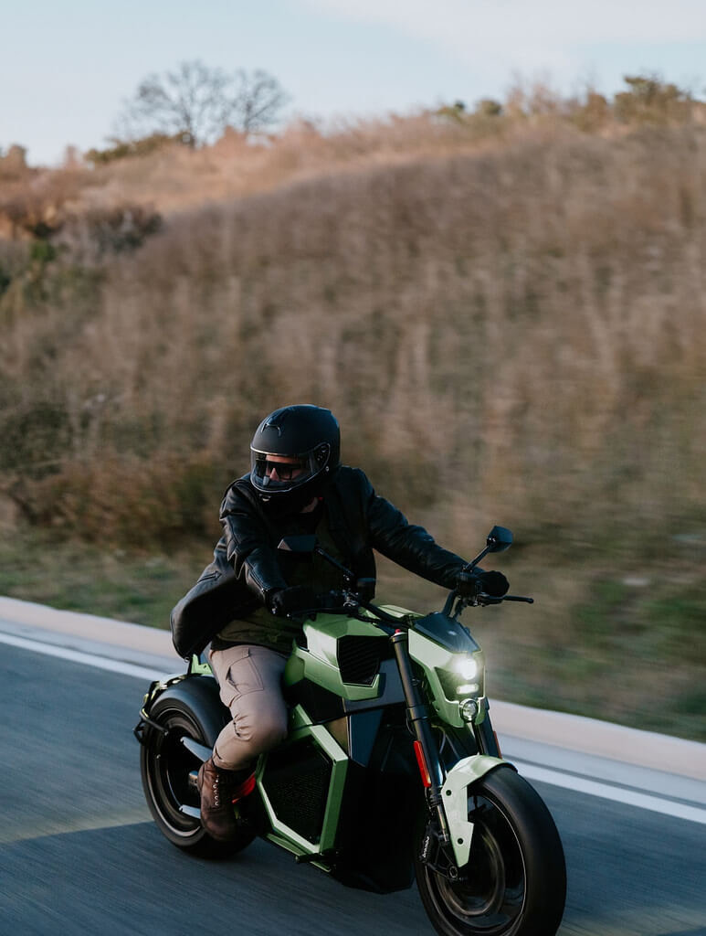 A close-up of a man driving an electric motorcycle. The driver wears a helmet, a black leather jacket and cargo pants. The motorcycle lights are on, as it is at twilight.