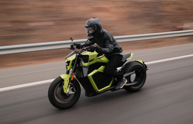 study-mob A person wearing a black helmet and jacket rides a bright green motorcycle at speed on a road. The background appears blurred, indicating motion. The road is bordered by a metal guardrail.