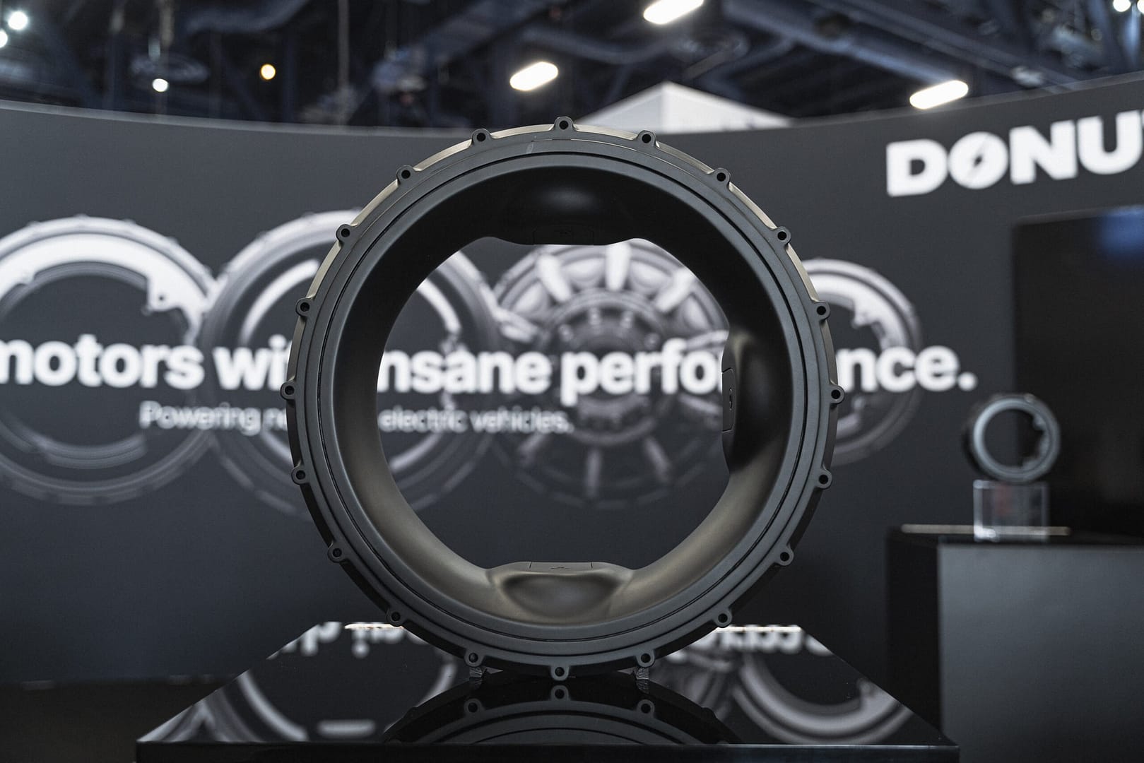 A circular motor component displayed on a stand at an exhibition. The background features partially visible text and another similar component. The lighting highlights the details and texture of the motor part.