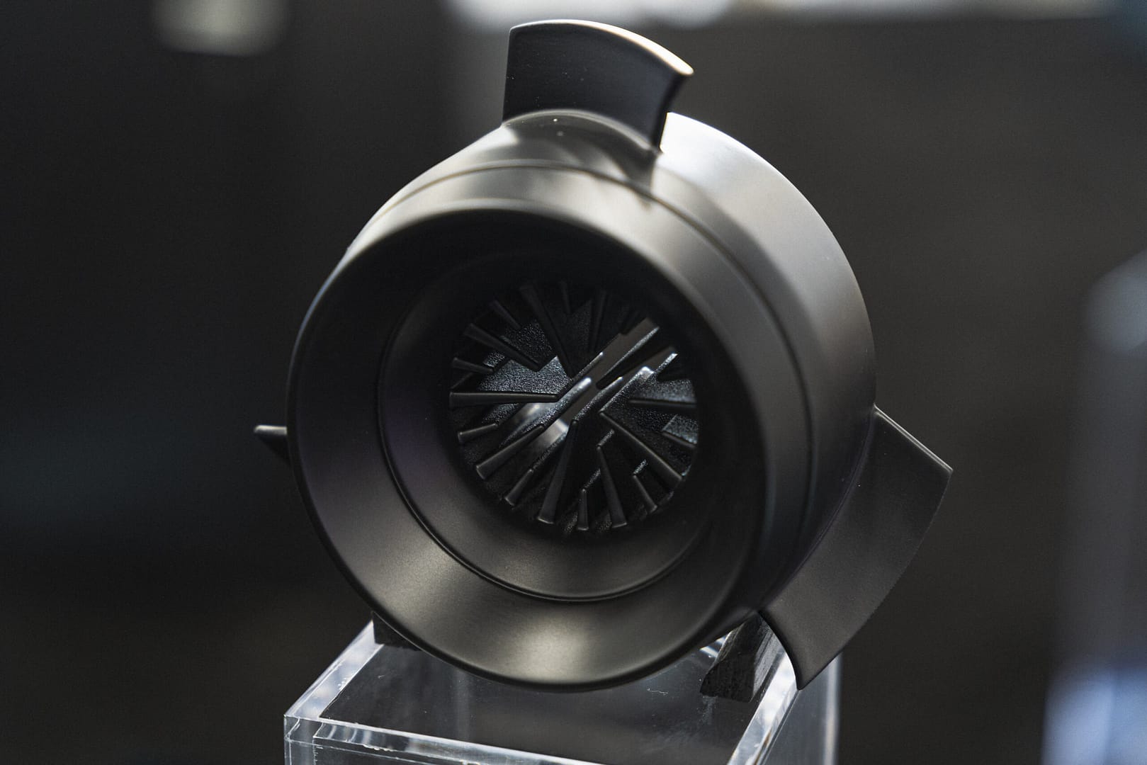 Close-up of a sleek, black, modern jet engine model displayed on a transparent stand. The design features intricate fan blades visible at the center, with a glossy and minimalist exterior. The background is blurred, emphasizing the model's details.