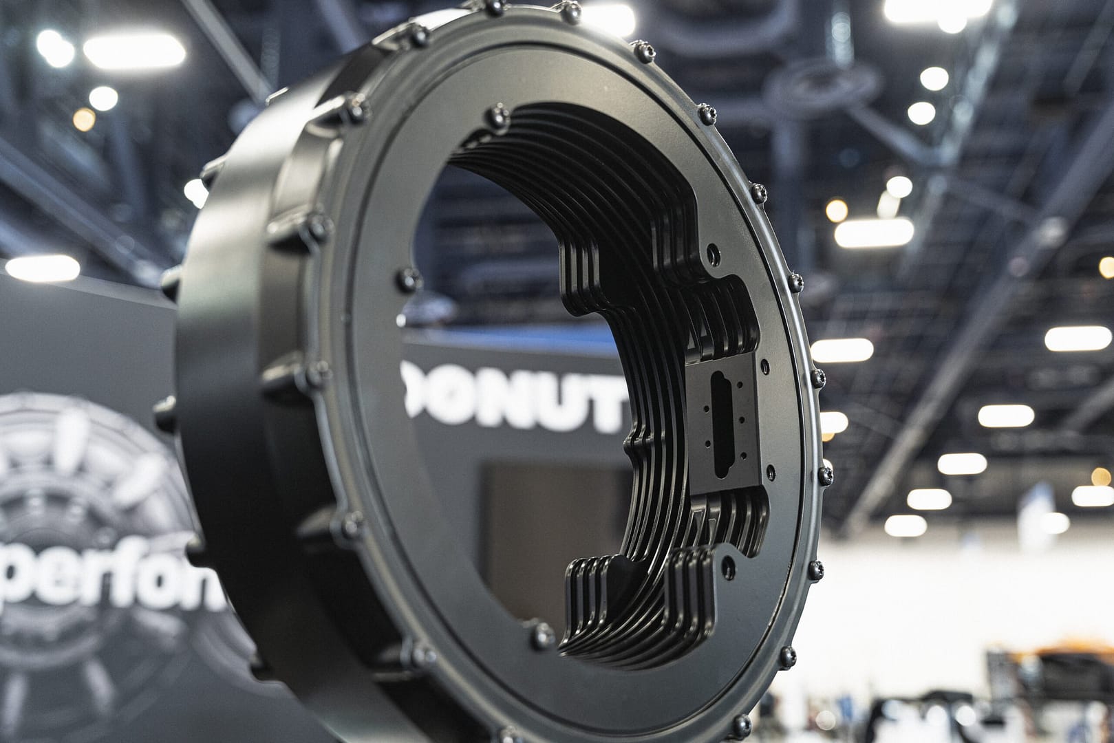 Close-up of a black metal automotive part with multiple bolts, displayed indoors. The background is blurred, featuring indistinct structures and lights, suggesting an exhibition or trade show setting.