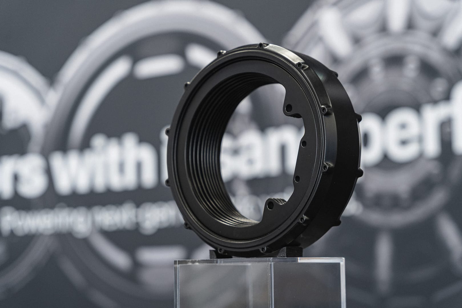 A black, cylindrical mechanical part with a threaded interior is displayed on a clear stand. The background is blurred, showing partial text and circular designs on a gray surface.