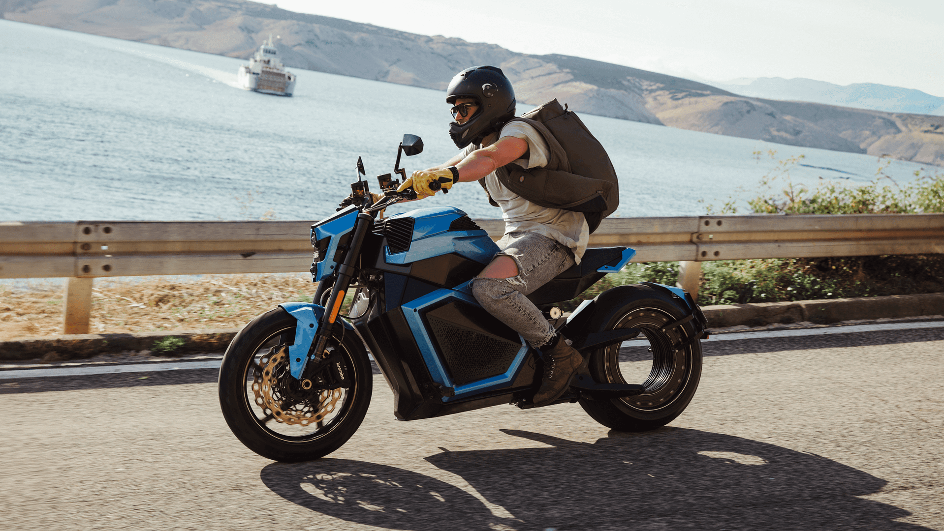 Rectangle 290 (1) A person wearing a helmet and backpack rides a futuristic blue motorcycle along a coastal road, with the ocean and a boat in the background. The scene is bright and sunny.
