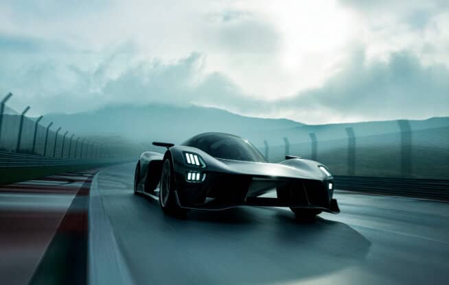 A sleek, futuristic car speeds on a race track under a cloudy sky. The vehicle's design features sharp angles and bright headlights. The surrounding environment appears wet, with mountains visible in the background.