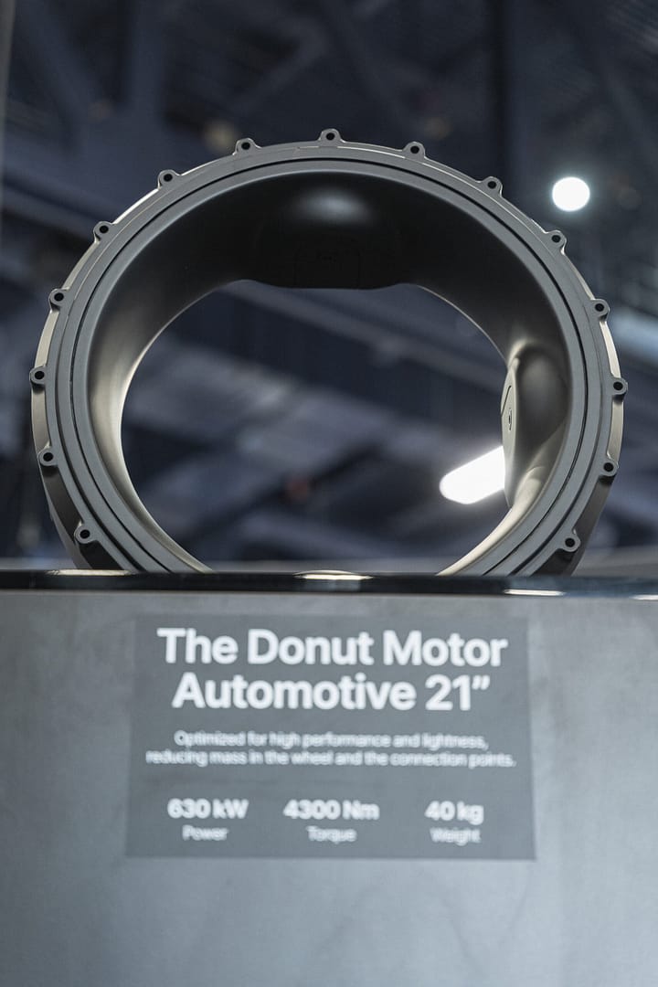 Close-up of a large, circular automotive component named "The Donut Motor Automotive 21" on display. The sign below reads: "630 kW Power, 4300 Nm Torque, 40 kg Weight." The background is blurred with overhead lights.