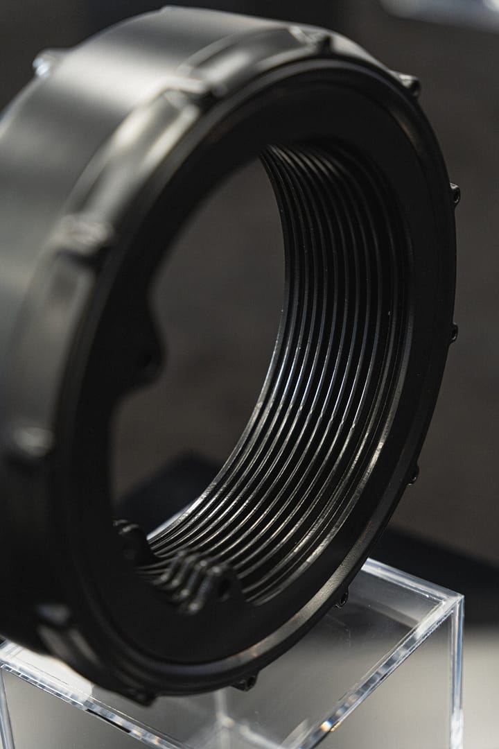 Close-up of a black, round, threaded metal or plastic component placed on a clear acrylic stand, against a blurred dark background. The object features intricate grooves and a shiny, reflective surface.