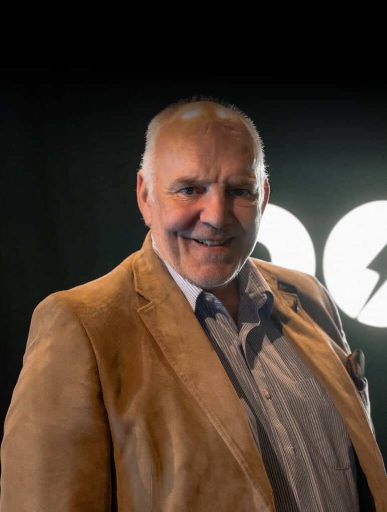 A man in a brown blazer and striped shirt stands smiling against a dark background with a white logo partially visible behind him.