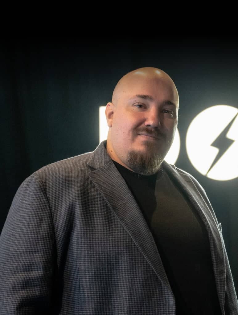 A person wearing a checkered blazer and black shirt stands confidently in front of a dark background with a logo that includes a lightning bolt.